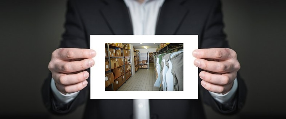 Eine Person, ohne Kopf, hält eine Karte in der Hand, auf der ein Lagerraum der Männerkammer zu sehen ist. Bestehend aus Regalen auf der linken Seite und Kleidersäcke auf der rechten Seite